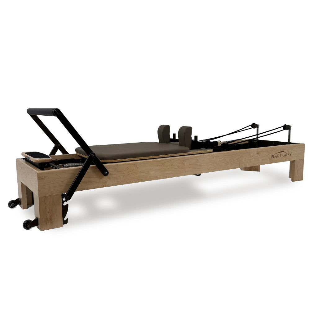 An Arbor™ reformer machine with a light natural wood frame, mocha brown padded carriage, and black metal handles and springs, shown on a clean white background.  The brand name 'PEAK PILATES®' is printed on the side of the frame.