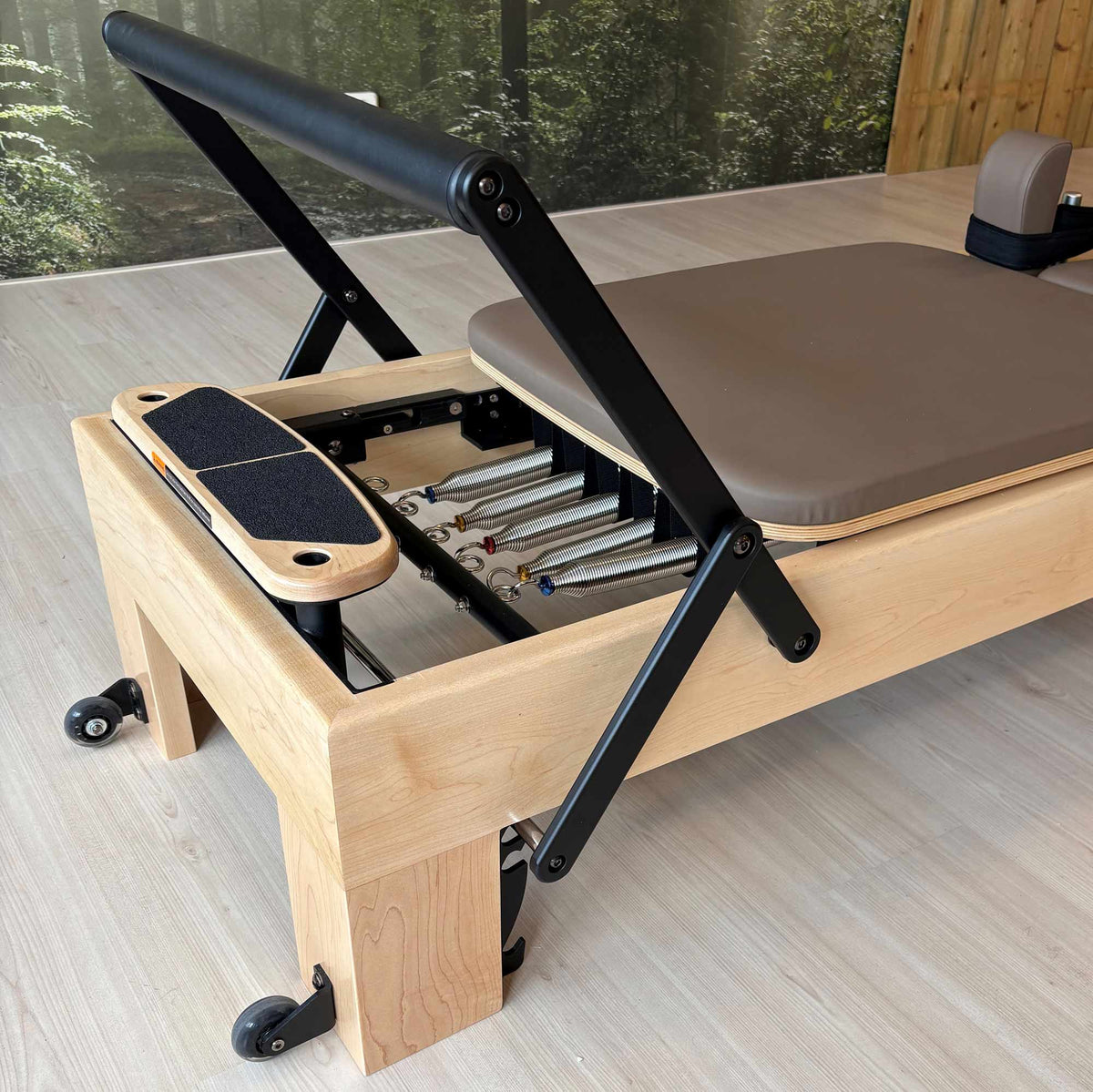 A Peak Pilates® reformer machine with black metal frame components, a mocha brown padded seat, and a foot platform, photographed in an indoor studio setting with wood flooring and a forest mural in the background.