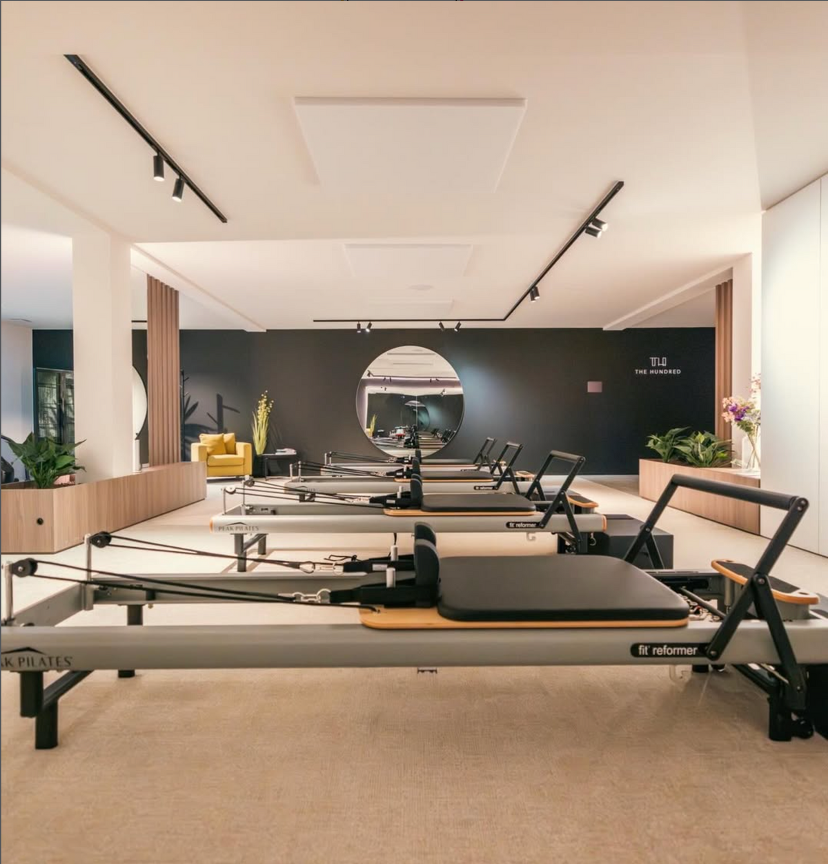 A modern Pilates reformer gym interior with multiple Peak Pilates reformer machines arranged in a spacious room with black accent walls, track lighting, and a large oval mirror.