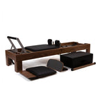 Pilates reformer exercise machine with dark wood frame, black cushioned carriage, metal guide bars, and several accessories including a black storage box, foam pad, resistance band, and wooden roller, photographed on a clean white studio background.