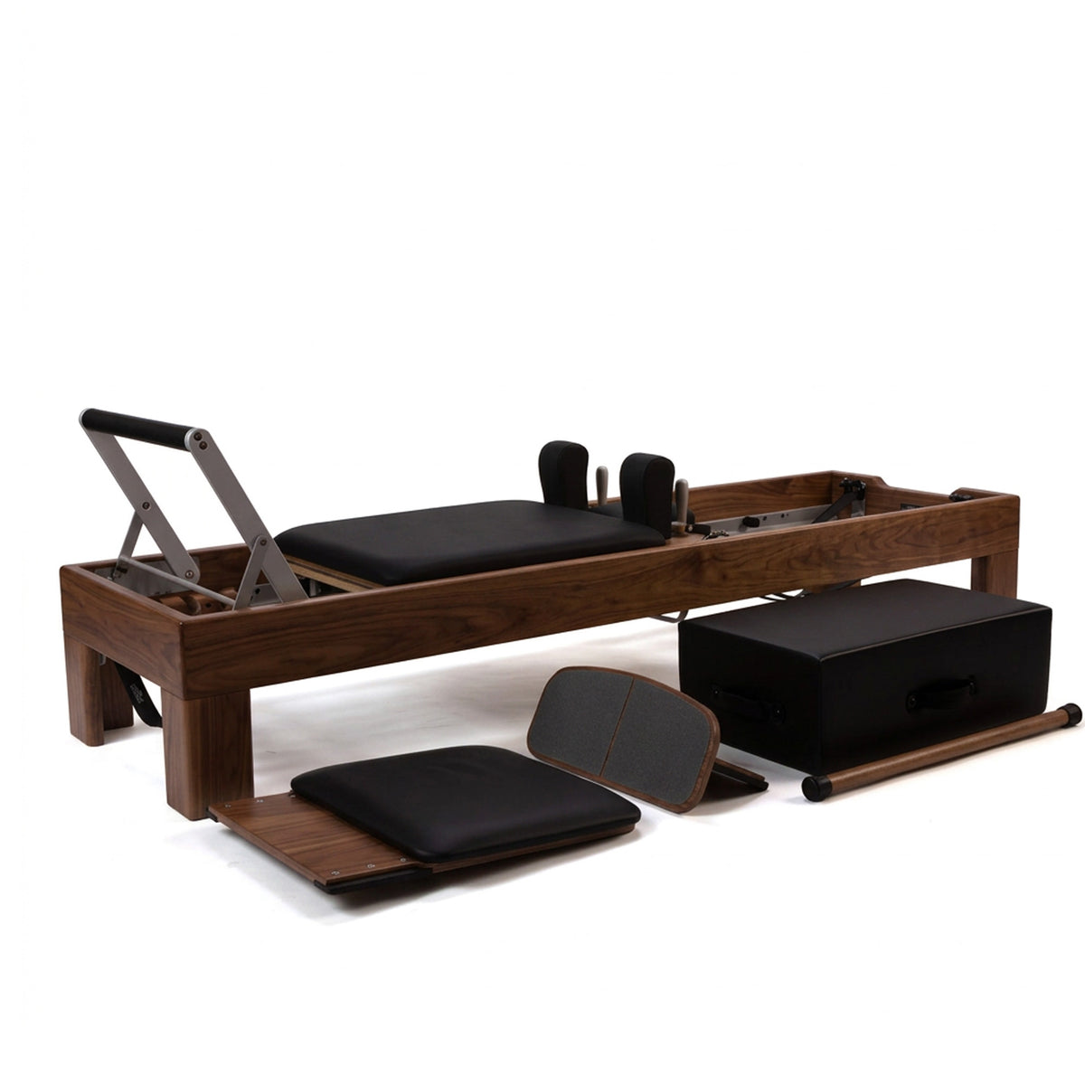 Pilates reformer exercise machine with dark wood frame, black cushioned carriage, metal guide bars, and several accessories including a black storage box, foam pad, resistance band, and wooden roller, photographed on a clean white studio background.